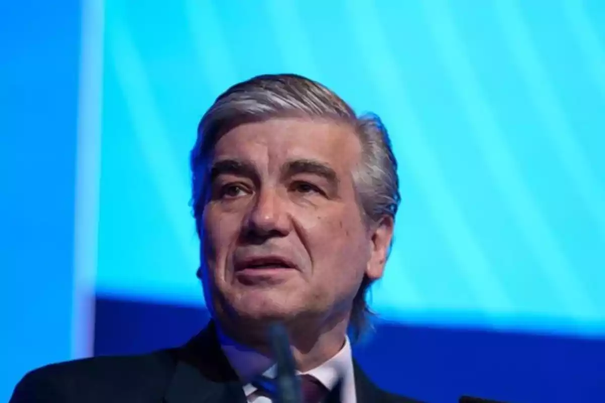 Hombre de cabello canoso y traje oscuro hablando frente a un fondo azul Hombre de cabello canoso y traje oscuro hablando frente a un fondo azul