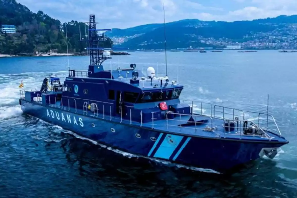 Barco azul de la aduana navegando en el mar cerca de la costa con montañas y una ciudad al fondo Barco azul de la aduana navegando en el mar cerca de la costa con montañas y una ciudad al fondo