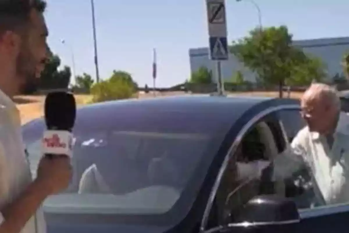 Un hombre sostiene un micrófono mientras otro hombre mayor se acerca a la puerta de un automóvil estacionado en la calle en un día soleado. Un hombre sostiene un micrófono mientras otro hombre mayor se acerca a la puerta de un automóvil estacionado en la calle en un día soleado.