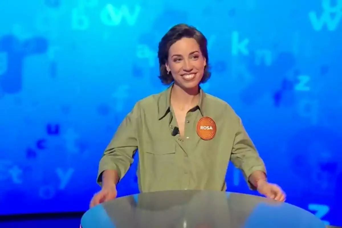 Rosa Rodríguez sonriente con camisa verde y un gafete naranja que dice Rosa participando en un programa de televisión, que es 'Pasapalabra', con fondo azul lleno de letras. Rosa Rodríguez sonriente con camisa verde y un gafete naranja que dice Rosa participando en un programa de televisión, que es 'Pasapalabra', con fondo azul lleno de letras.