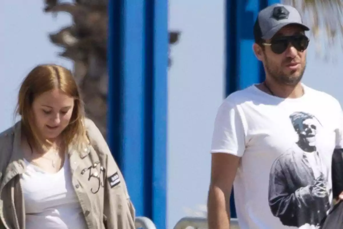 Dos personas caminando al aire libre en un día soleado, una de ellas lleva gafas de sol y gorra, la otra viste una chaqueta beige Dos personas caminando al aire libre en un día soleado, una de ellas lleva gafas de sol y gorra, la otra viste una chaqueta beige