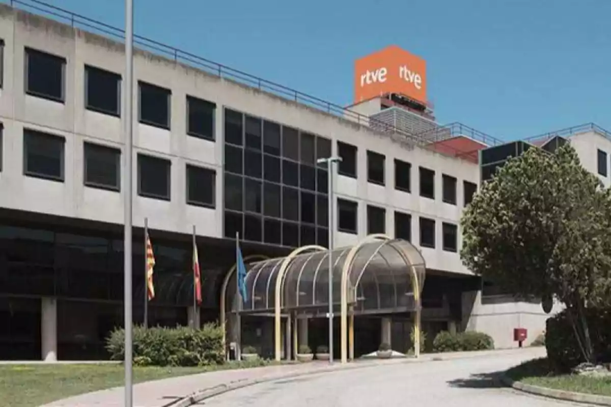 Edificio de RTVE con fachada de ventanas oscuras, entrada principal con techo curvo de cristal y varias banderas en el exterior Edificio de RTVE con fachada de ventanas oscuras, entrada principal con techo curvo de cristal y varias banderas en el exterior