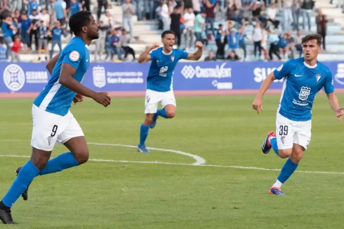 Tres futbolistas con uniforme azul celebran un gol en un estadio mientras el público aplaude en las gradas. Tres futbolistas con uniforme azul celebran un gol en un estadio mientras el público aplaude en las gradas.