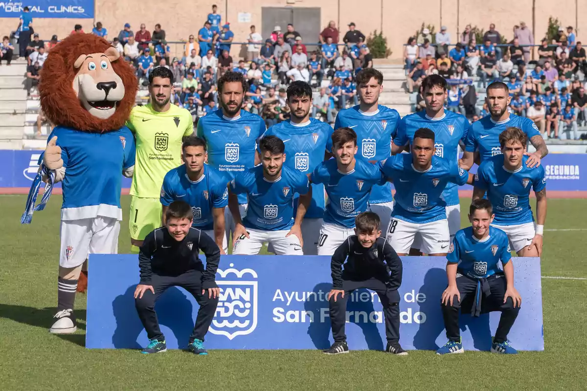 Equipo de fútbol posando para una foto grupal en el campo junto a su mascota y tres niños frente a una pancarta azul con público en las gradas al fondo Equipo de fútbol posando para una foto grupal en el campo junto a su mascota y tres niños frente a una pancarta azul con público en las gradas al fondo