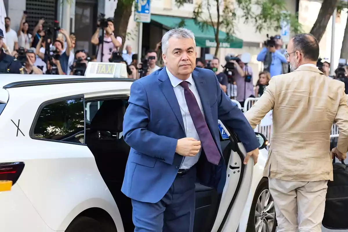 Hombre con traje azul saliendo de un taxi blanco mientras varias personas y fotógrafos lo observan en la calle Hombre con traje azul saliendo de un taxi blanco mientras varias personas y fotógrafos lo observan en la calle
