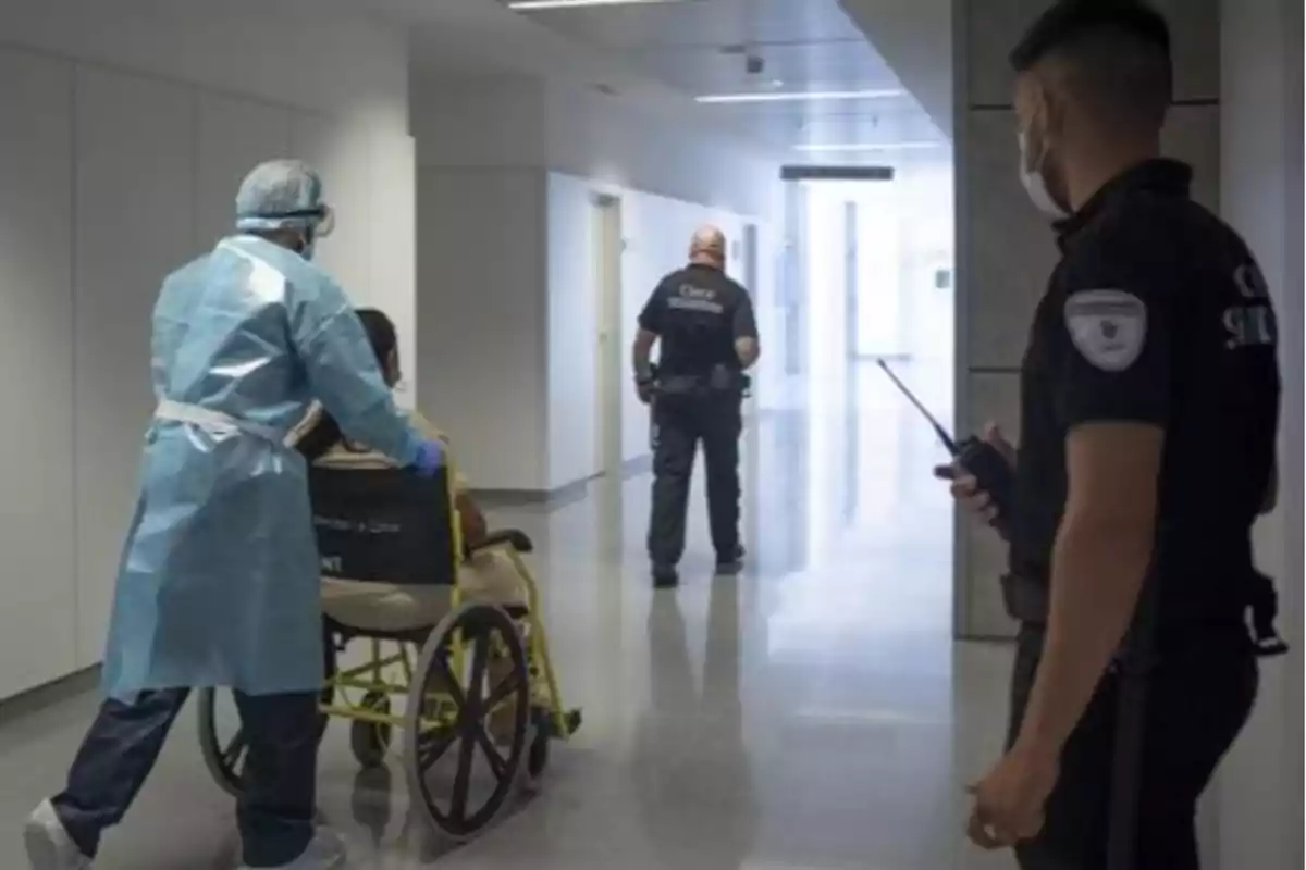 Un trabajador de la salud empuja una silla de ruedas por un pasillo de hospital mientras dos guardias de seguridad observan