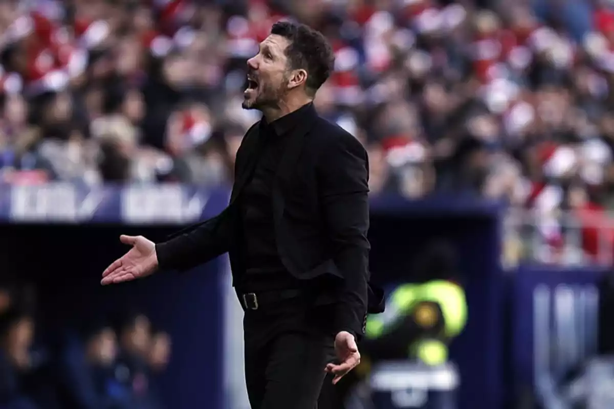 Entrenador de fútbol vestido de negro gesticulando y gritando en la banda durante un partido con el público de fondo