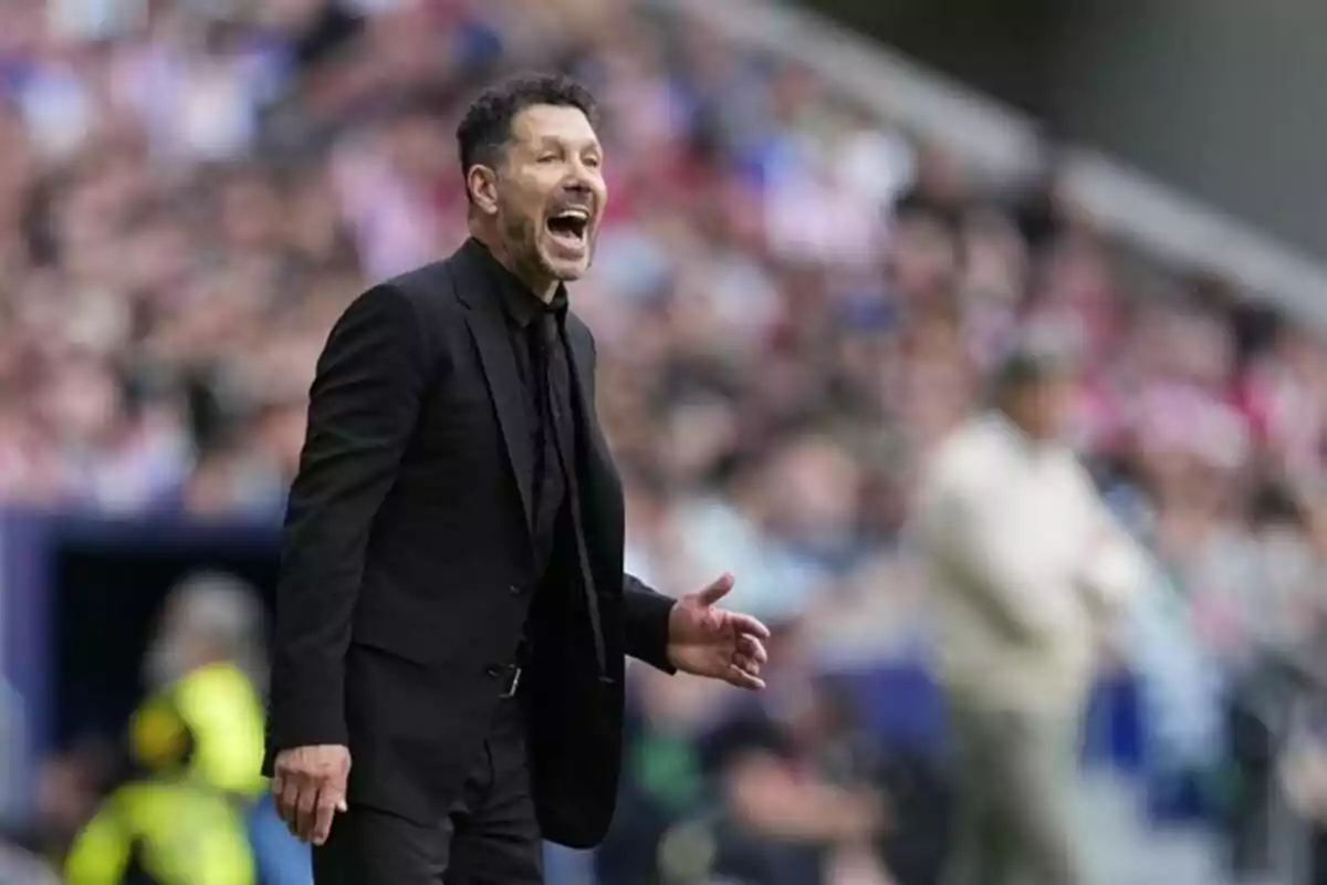 Entrenador de fútbol vestido de negro dando indicaciones en la banda durante un partido con el público desenfocado al fondo Entrenador de fútbol vestido de negro dando indicaciones en la banda durante un partido con el público desenfocado al fondo