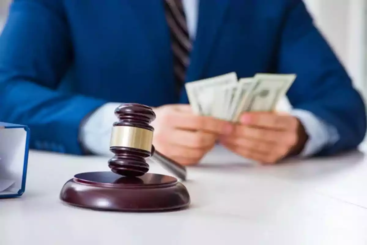Persona con traje azul contando billetes de dinero junto a un mazo de juez sobre una mesa Persona con traje azul contando billetes de dinero junto a un mazo de juez sobre una mesa