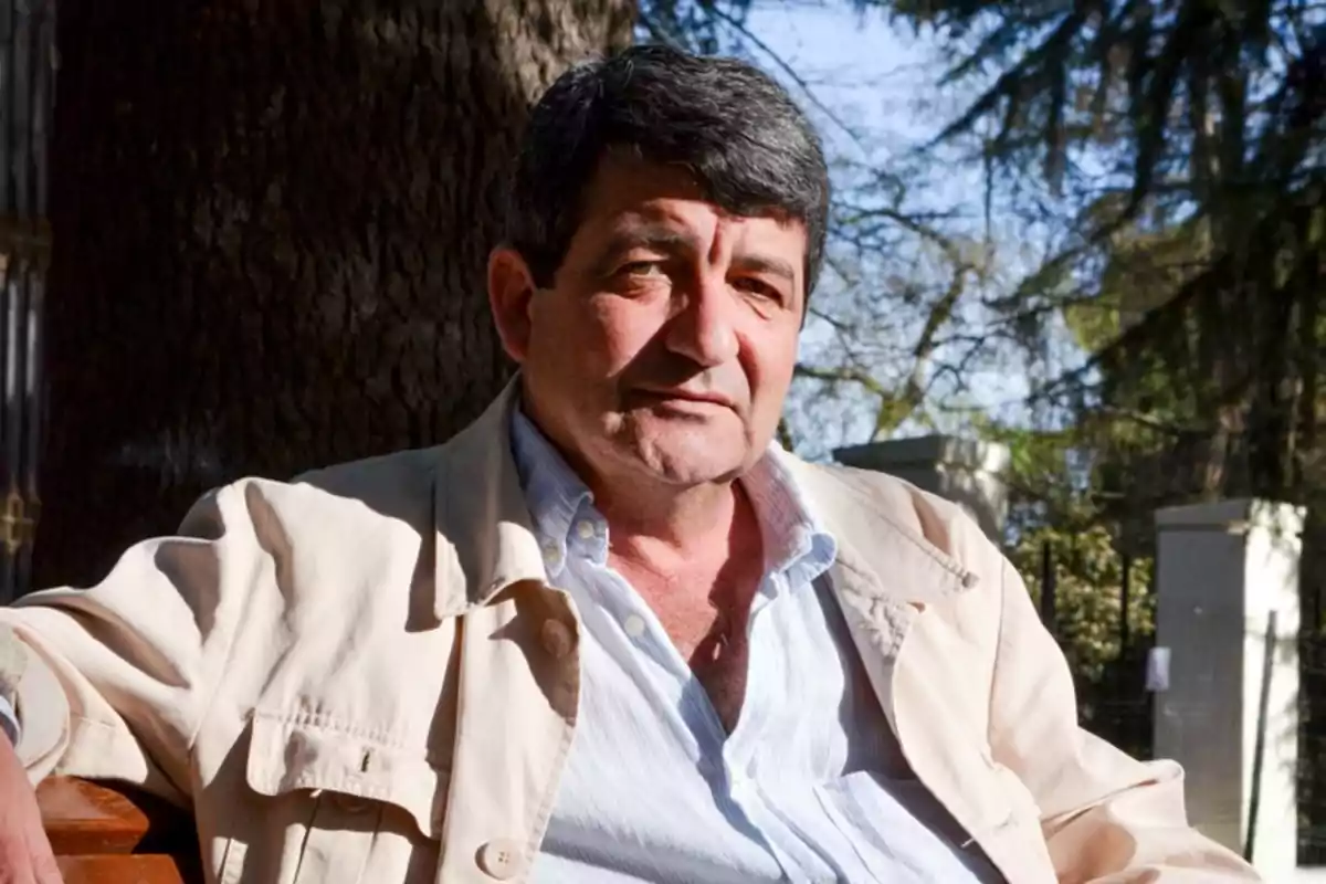Hombre de cabello canoso y corto sentado al aire libre con árboles y una cerca al fondo
