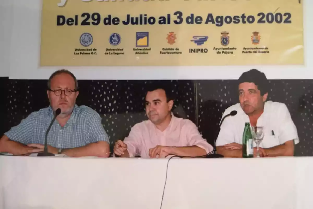 Tres hombres sentados en una mesa durante una conferencia con un cartel amarillo de fondo que indica fechas y nombres de instituciones organizadoras del evento