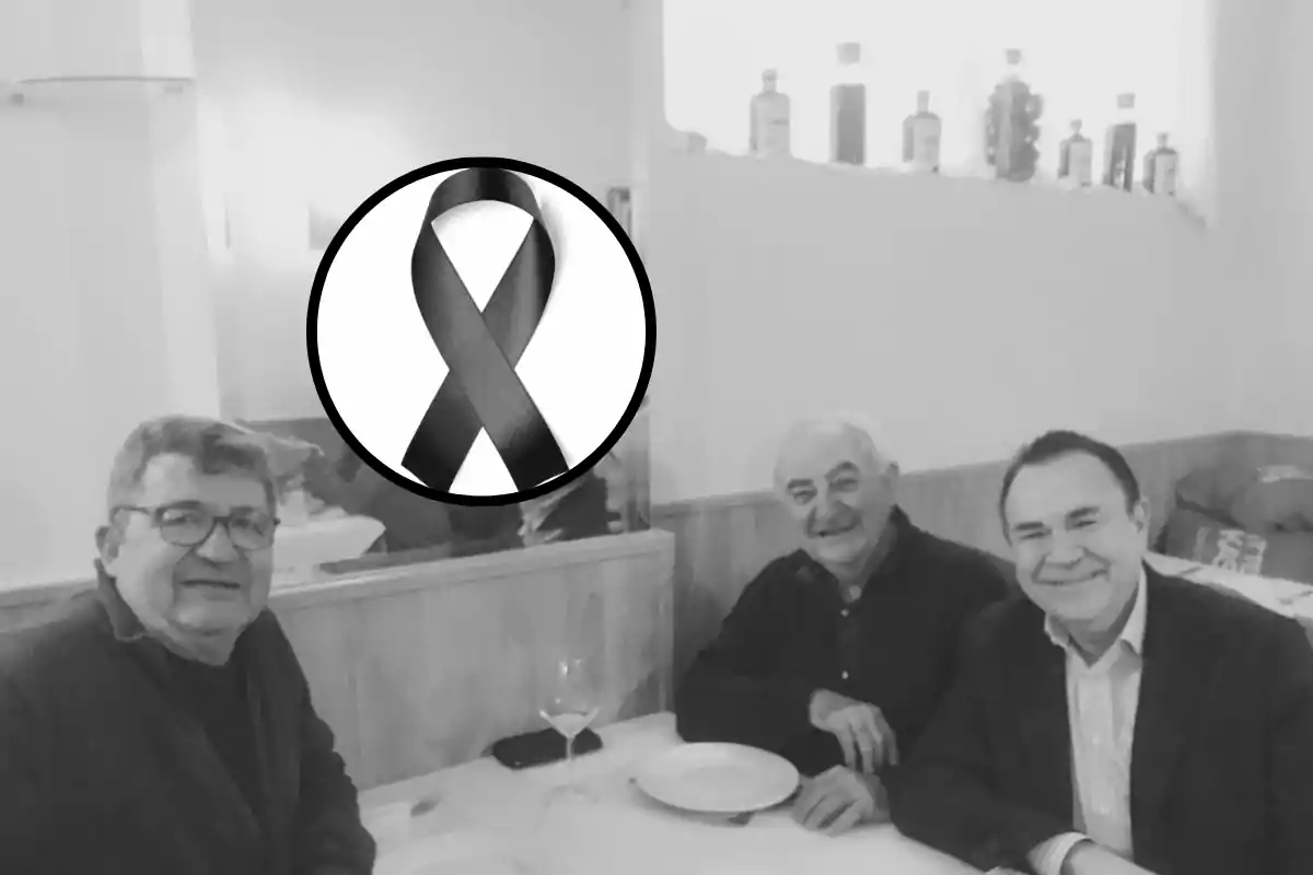 Tres hombres mayores sentados en una mesa de restaurante con platos y copas vacías, la imagen está en blanco y negro y tiene un lazo negro de luto en la parte superior izquierda