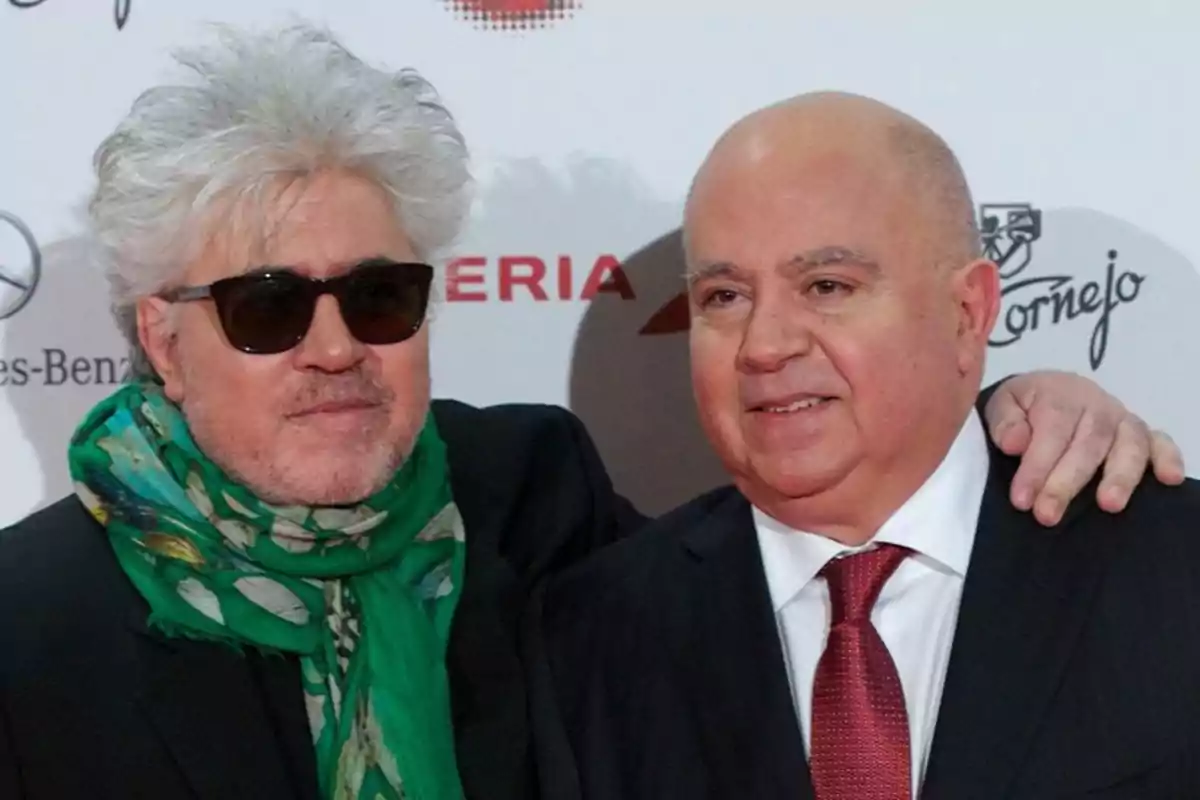Dos hombres posan juntos en un evento formal uno lleva gafas de sol y bufanda verde mientras el otro viste traje y corbata roja