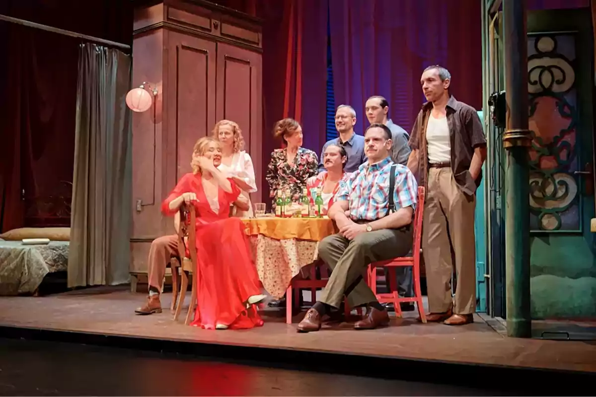 Un grupo de actores en el escenario interpreta una escena alrededor de una mesa con botellas y vasos, en un ambiente que parece ser una casa antigua decorada con cortinas rojas y muebles clásicos