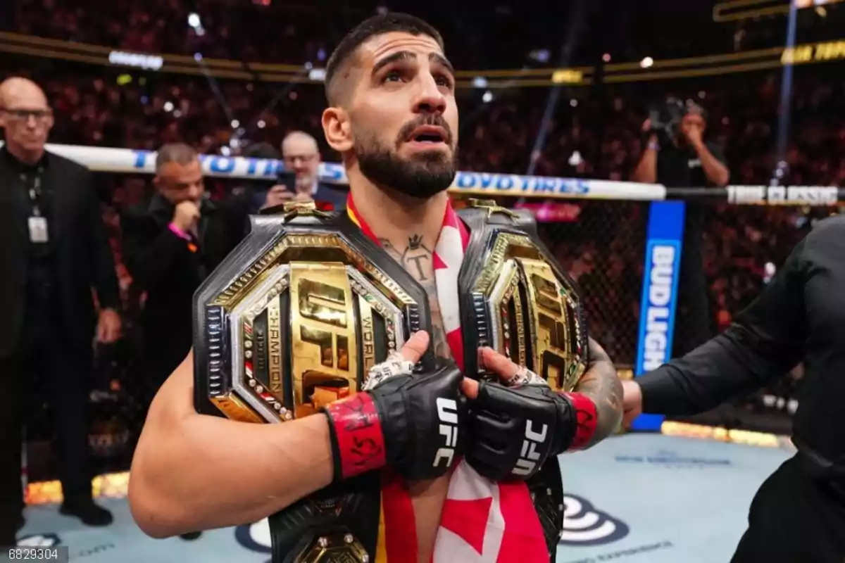 Un peleador de artes marciales mixtas sostiene dos cinturones de campeón de la UFC dentro del octágono mientras lleva una bandera sobre los hombros