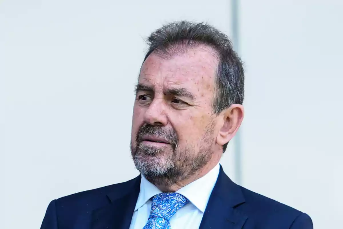Hombre de mediana edad con barba y cabello corto, vestido con traje azul y corbata estampada, mirando hacia un lado sobre un fondo claro Hombre de mediana edad con barba y cabello corto, vestido con traje azul y corbata estampada, mirando hacia un lado sobre un fondo claro