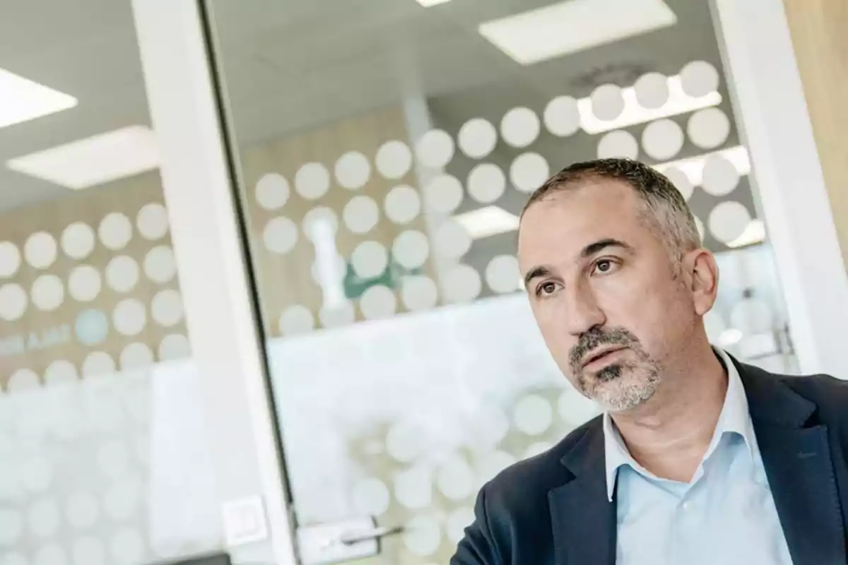Hombre de mediana edad con barba corta y cabello corto, vestido con saco azul y camisa celeste, sentado en una oficina moderna con paredes de vidrio decoradas con círculos blancos Hombre de mediana edad con barba corta y cabello corto, vestido con saco azul y camisa celeste, sentado en una oficina moderna con paredes de vidrio decoradas con círculos blancos
