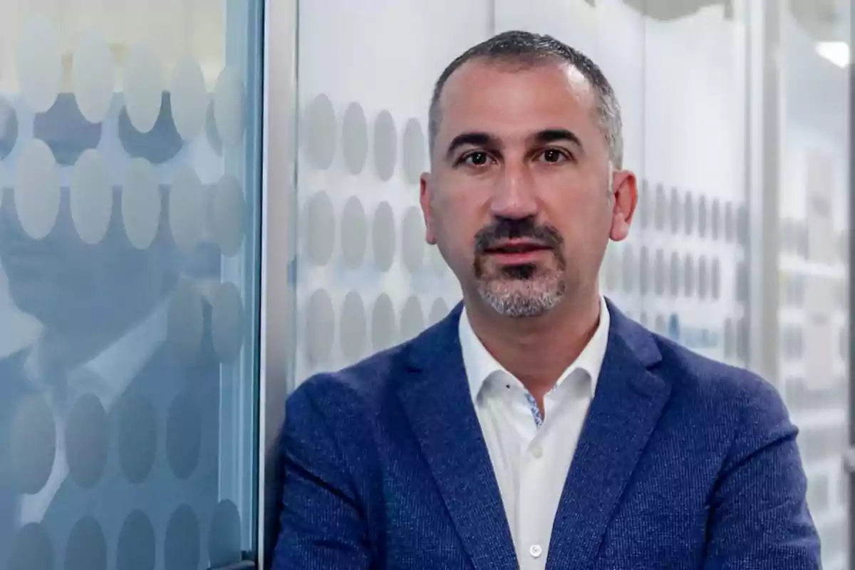 Hombre de cabello corto y barba de candado con saco azul y camisa blanca posando junto a una pared de vidrio con círculos translúcidos Hombre de cabello corto y barba de candado con saco azul y camisa blanca posando junto a una pared de vidrio con círculos translúcidos