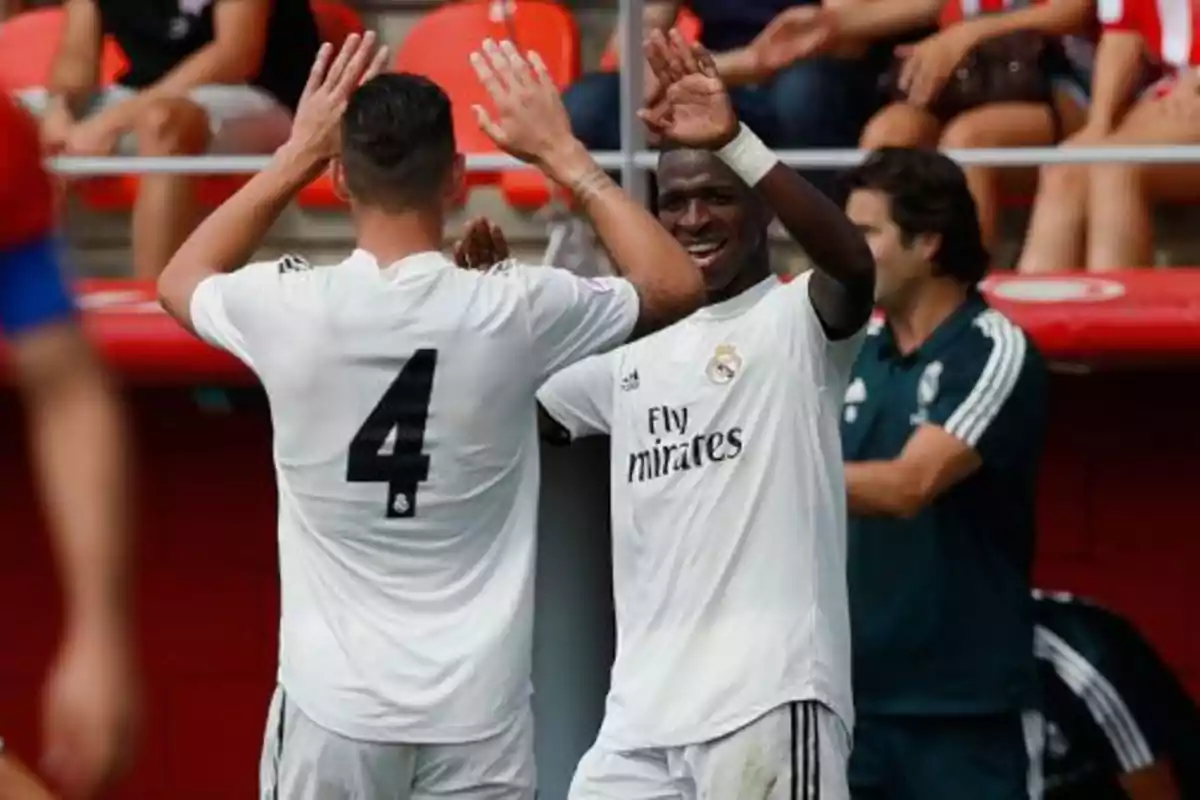 Dos futbolistas del Real Madrid celebran con un choque de manos durante un partido, ambos visten uniforme blanco y están sonrientes en el campo Dos futbolistas del Real Madrid celebran con un choque de manos durante un partido, ambos visten uniforme blanco y están sonrientes en el campo