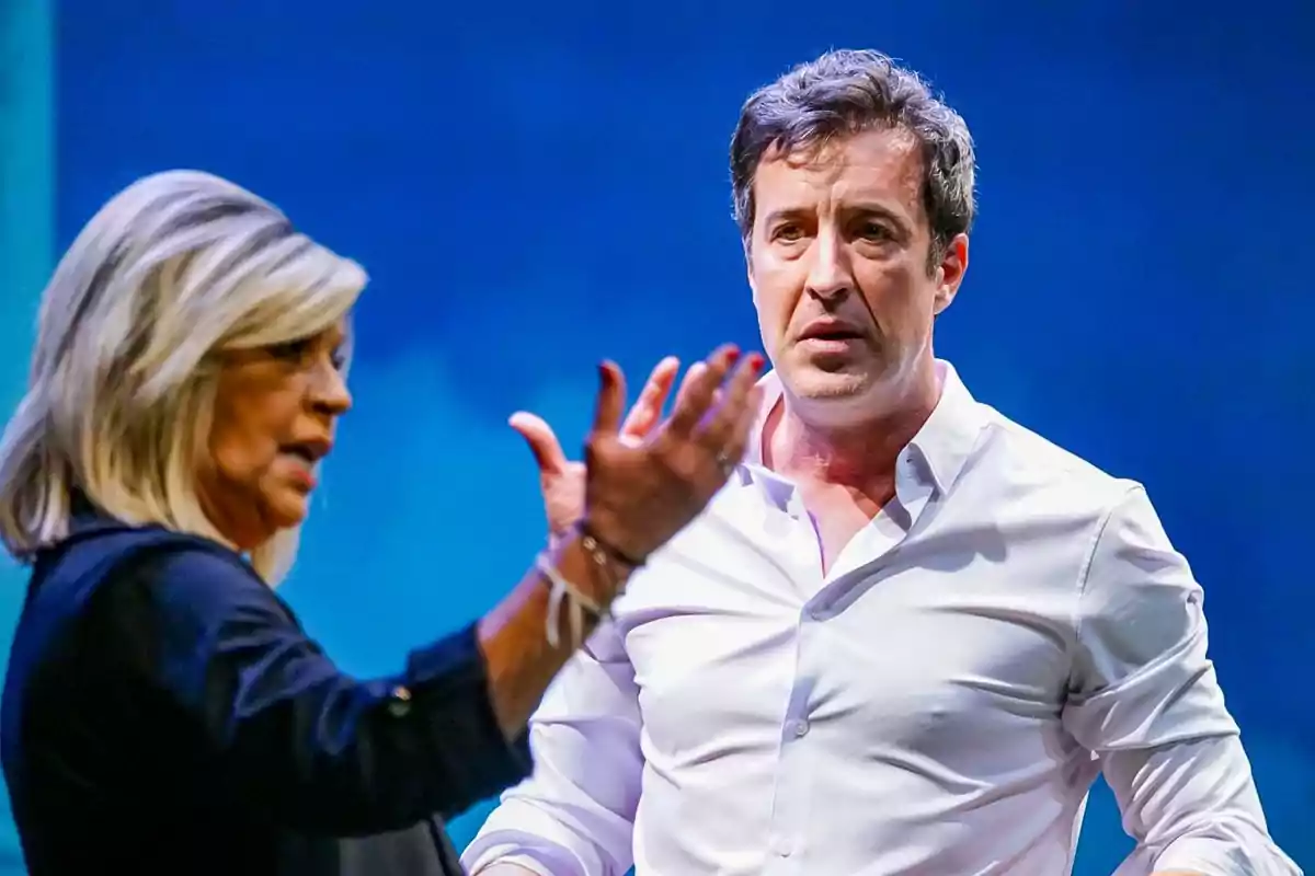 Dos personas conversan en un escenario con fondo azul, una mujer gesticula con la mano mientras un hombre la observa con expresión seria. Dos personas conversan en un escenario con fondo azul, una mujer gesticula con la mano mientras un hombre la observa con expresión seria.