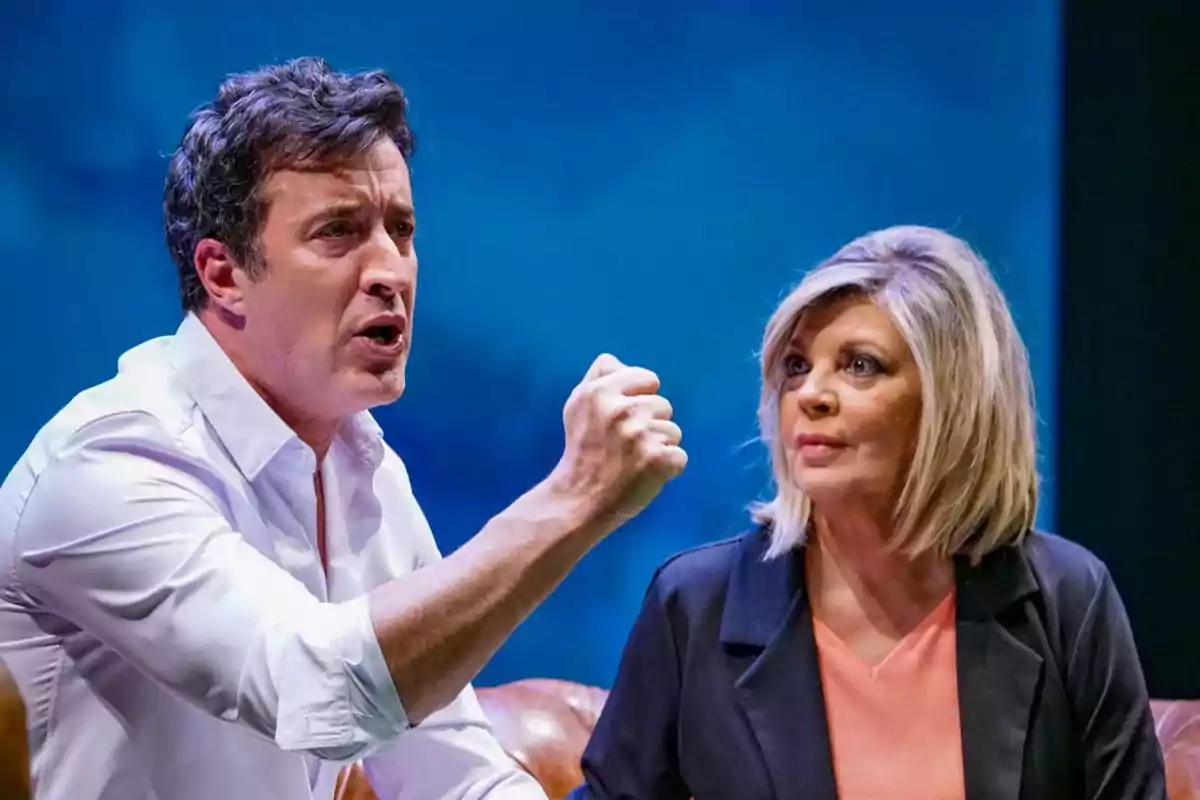 Hombre con camisa blanca gesticulando con el puño levantado mientras una mujer rubia lo observa atentamente en un escenario teatral