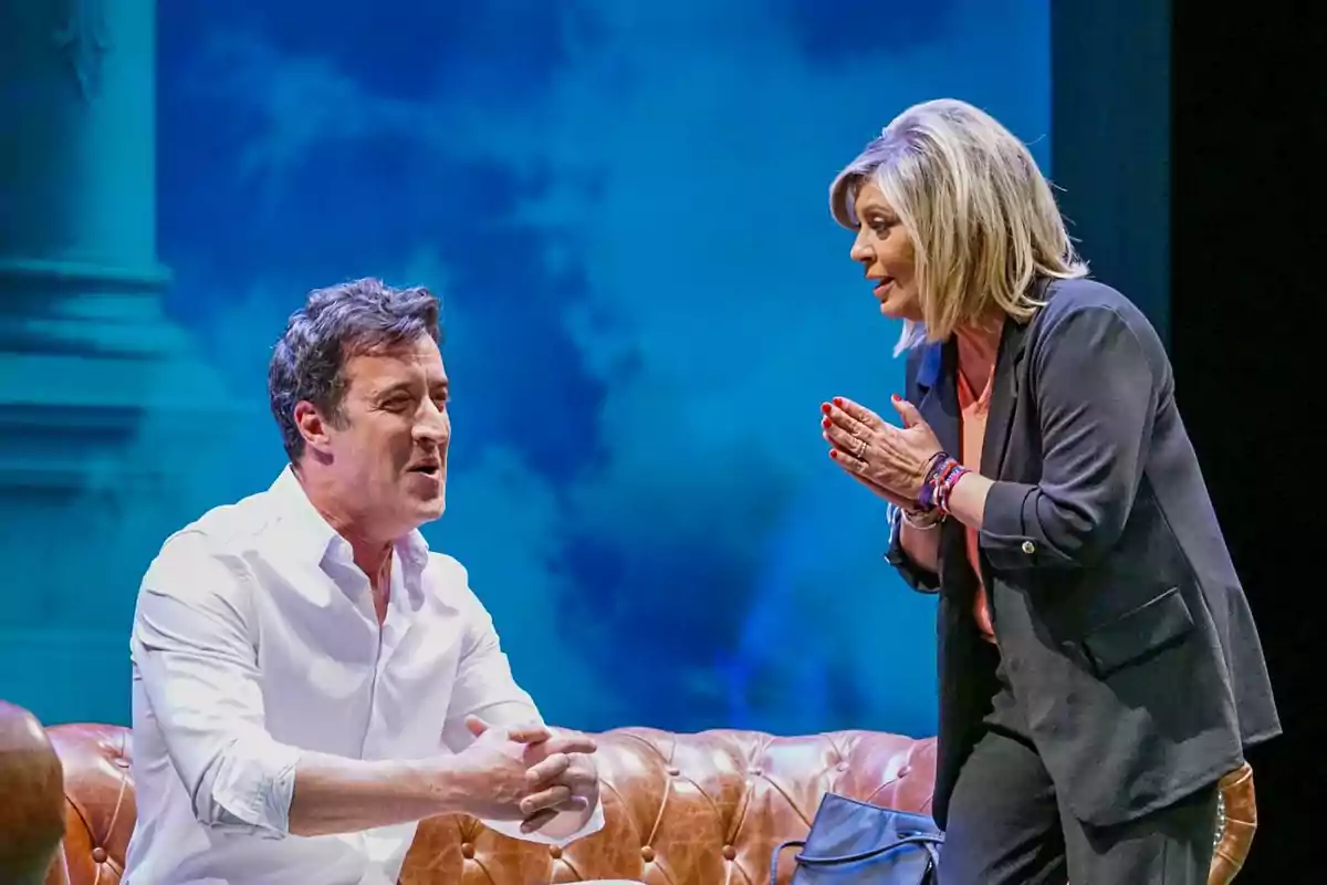 Dos personas conversan intensamente sobre un sofá de cuero en un escenario teatral con fondo azul Dos personas conversan intensamente sobre un sofá de cuero en un escenario teatral con fondo azul