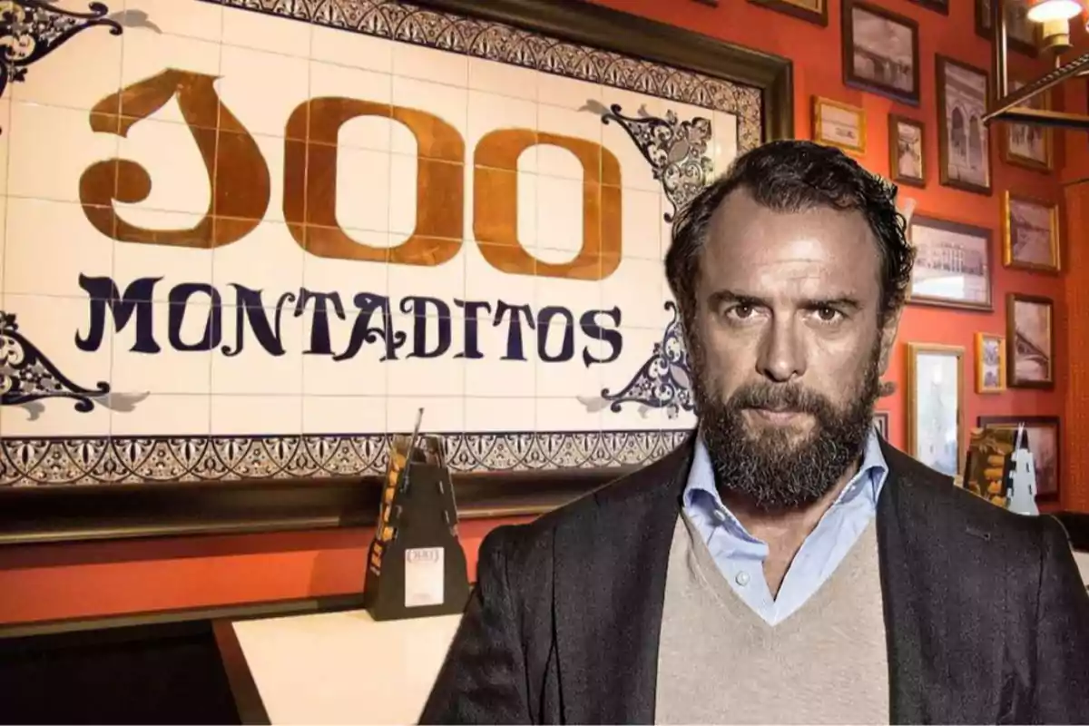 Hombre de barba y cabello oscuro con expresión seria posando frente a un letrero de 100 Montaditos en el interior de un restaurante decorado con cuadros en la pared