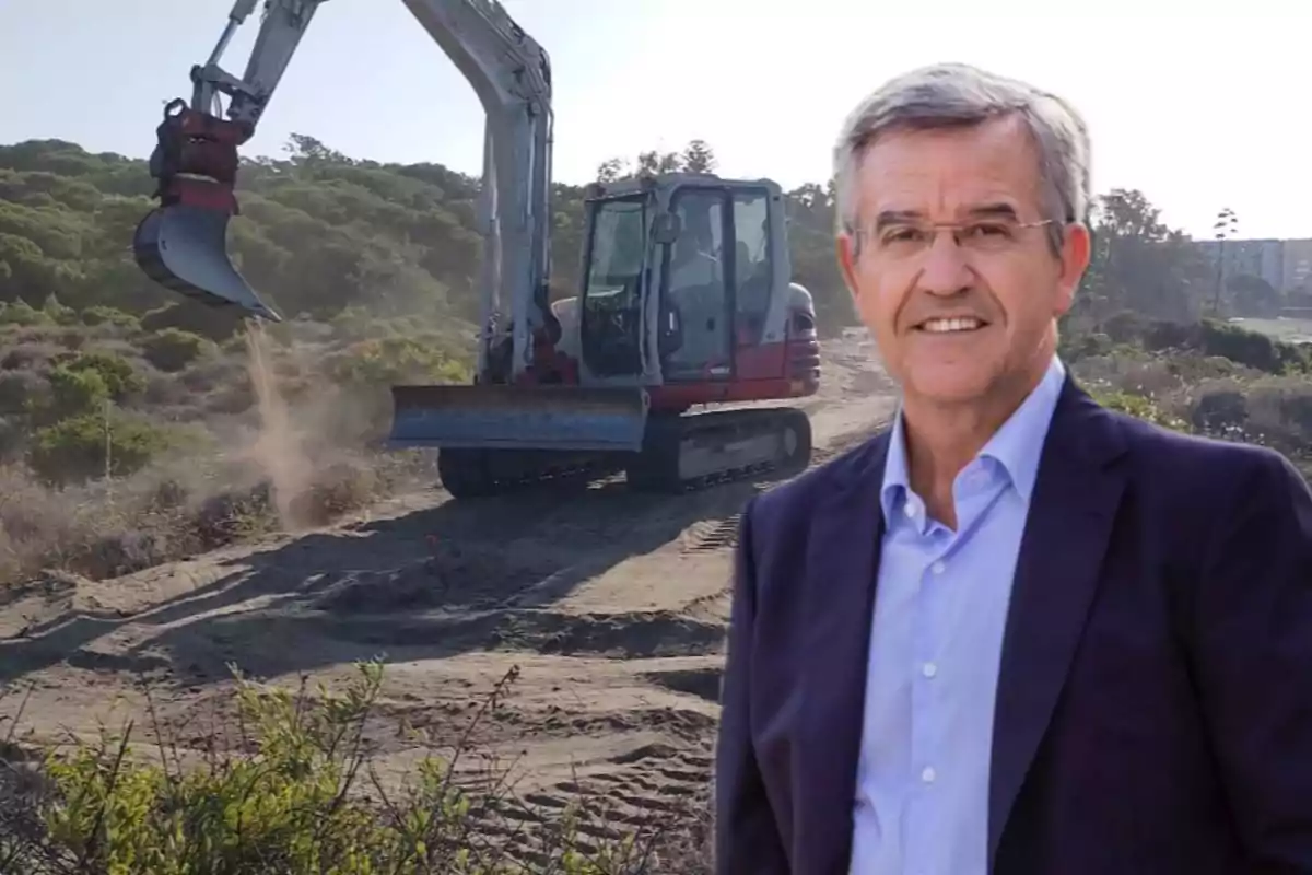Hombre de cabello canoso con gafas y saco azul claro sonríe frente a una excavadora trabajando en un terreno con vegetación y árboles al fondo