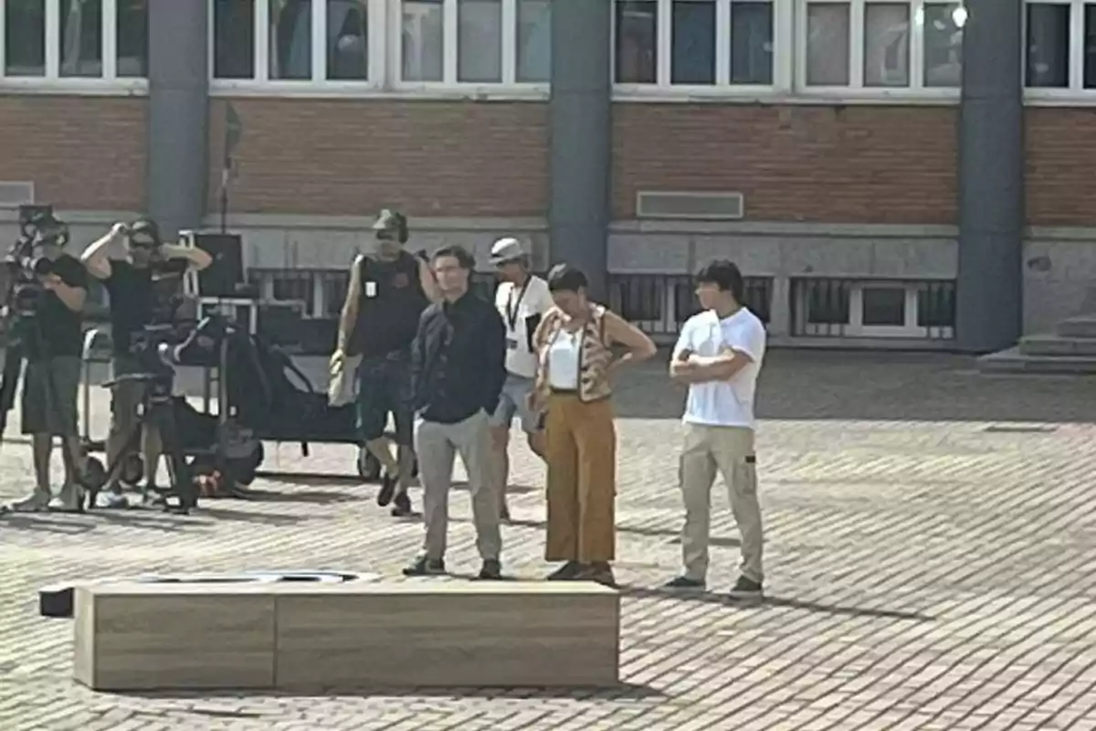 Varias personas se encuentran reunidas en una plaza frente a un edificio, algunas parecen estar trabajando con equipo de grabación y otras están de pie conversando. Varias personas se encuentran reunidas en una plaza frente a un edificio, algunas parecen estar trabajando con equipo de grabación y otras están de pie conversando.