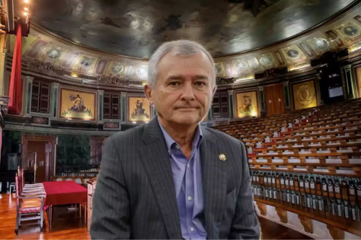 Hombre de cabello canoso con saco gris y camisa azul posando en el interior de un auditorio antiguo con asientos de madera y decoraciones clásicas en las paredes.