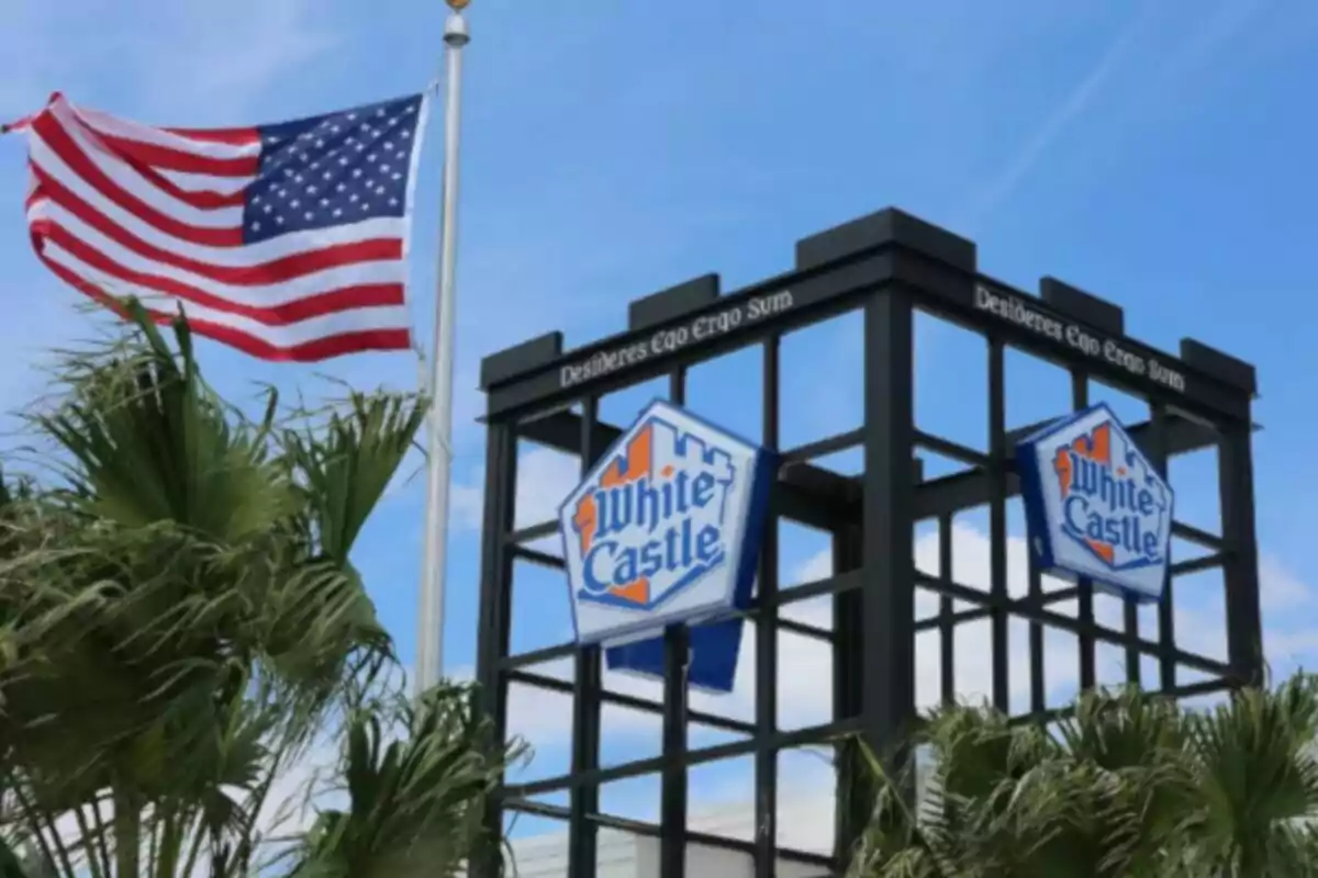 Bandera de Estados Unidos ondeando junto a un letrero de White Castle rodeado de palmeras bajo un cielo azul Bandera de Estados Unidos ondeando junto a un letrero de White Castle rodeado de palmeras bajo un cielo azul