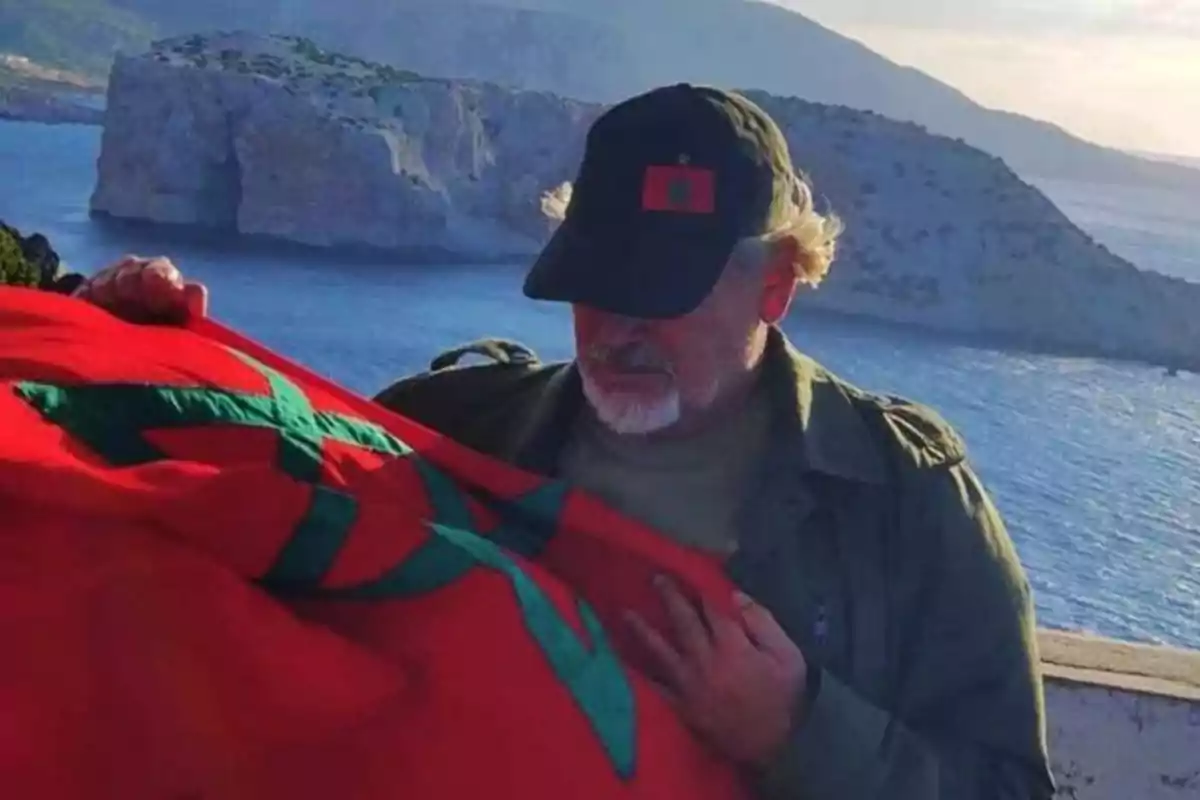 Hombre con gorra y chaqueta sosteniendo una bandera de Marruecos frente al mar con acantilados al fondo Hombre con gorra y chaqueta sosteniendo una bandera de Marruecos frente al mar con acantilados al fondo