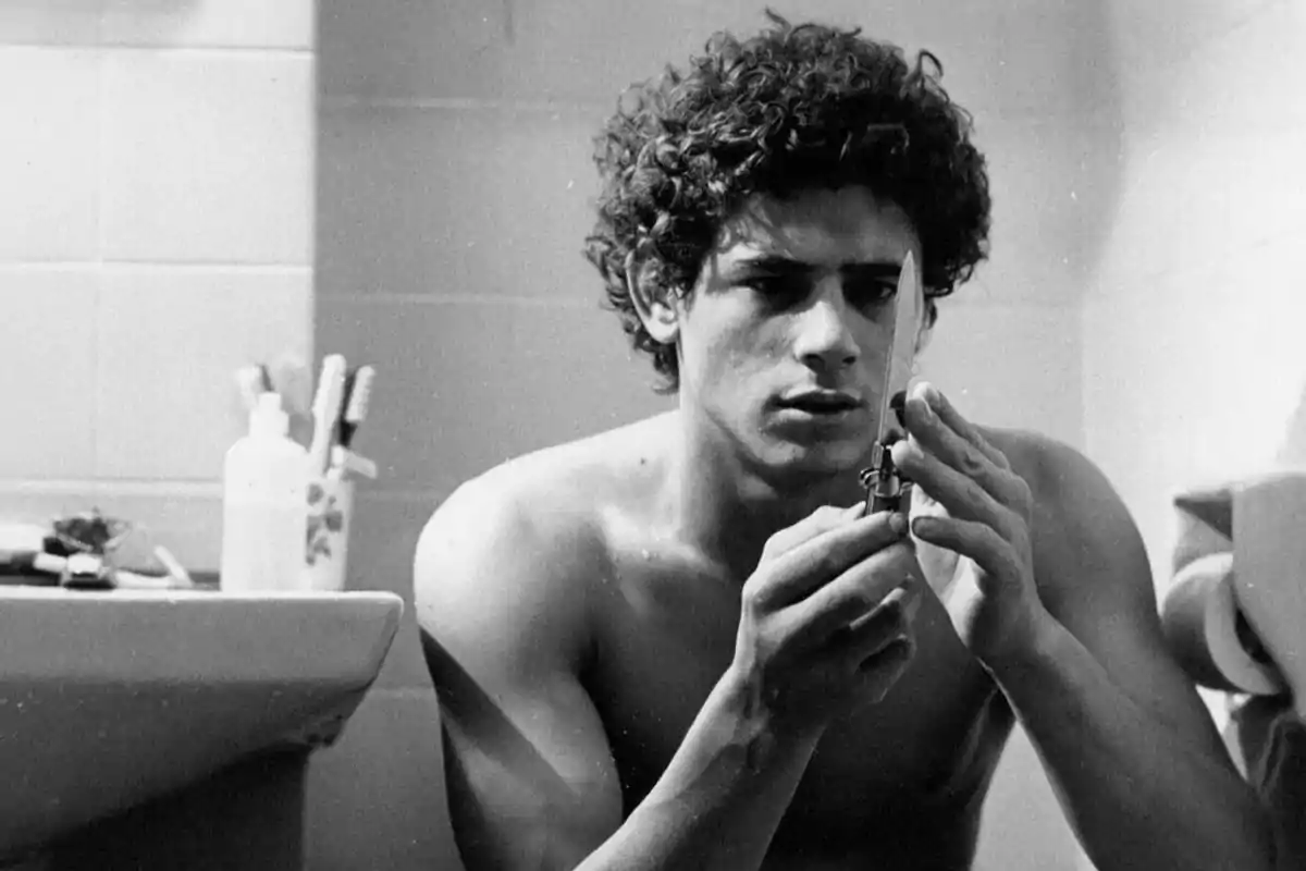 Joven de cabello rizado y torso desnudo sentado en un baño observando atentamente una navaja. Joven de cabello rizado y torso desnudo sentado en un baño observando atentamente una navaja.