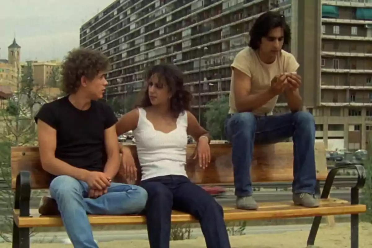 Tres jóvenes sentados en una banca conversan al aire libre con un edificio grande de fondo Tres jóvenes sentados en una banca conversan al aire libre con un edificio grande de fondo