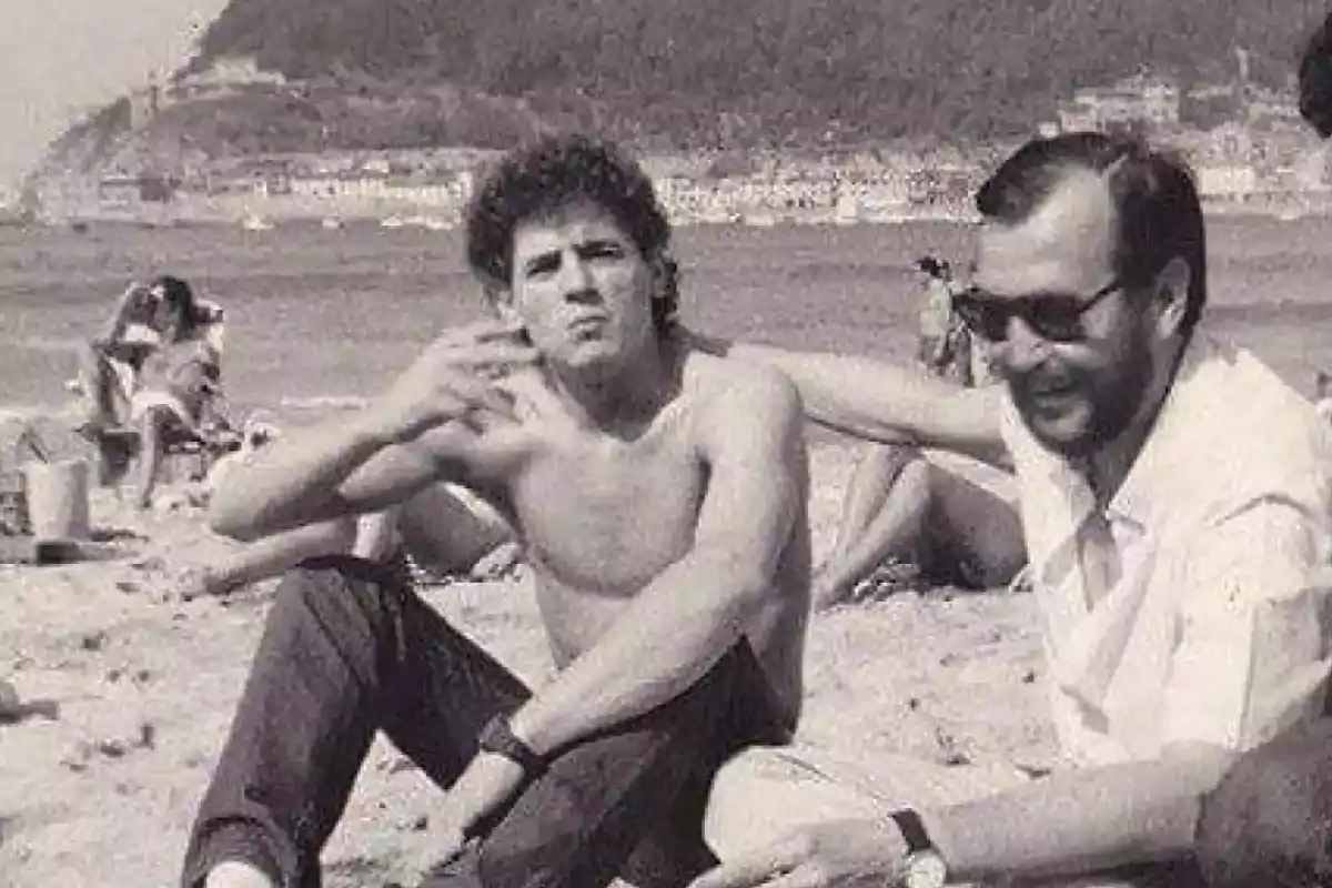 Dos hombres sentados en la playa uno de ellos sin camisa y el otro con gafas de sol ambos parecen relajados y al fondo se ve el mar y algunas personas disfrutando del día Dos hombres sentados en la playa uno de ellos sin camisa y el otro con gafas de sol ambos parecen relajados y al fondo se ve el mar y algunas personas disfrutando del día