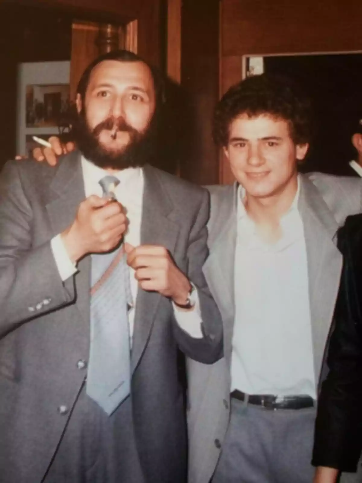 Dos hombres vestidos con traje posan juntos en un ambiente interior uno de ellos sostiene un cigarro y un encendedor mientras sonríen a la cámara Dos hombres vestidos con traje posan juntos en un ambiente interior uno de ellos sostiene un cigarro y un encendedor mientras sonríen a la cámara