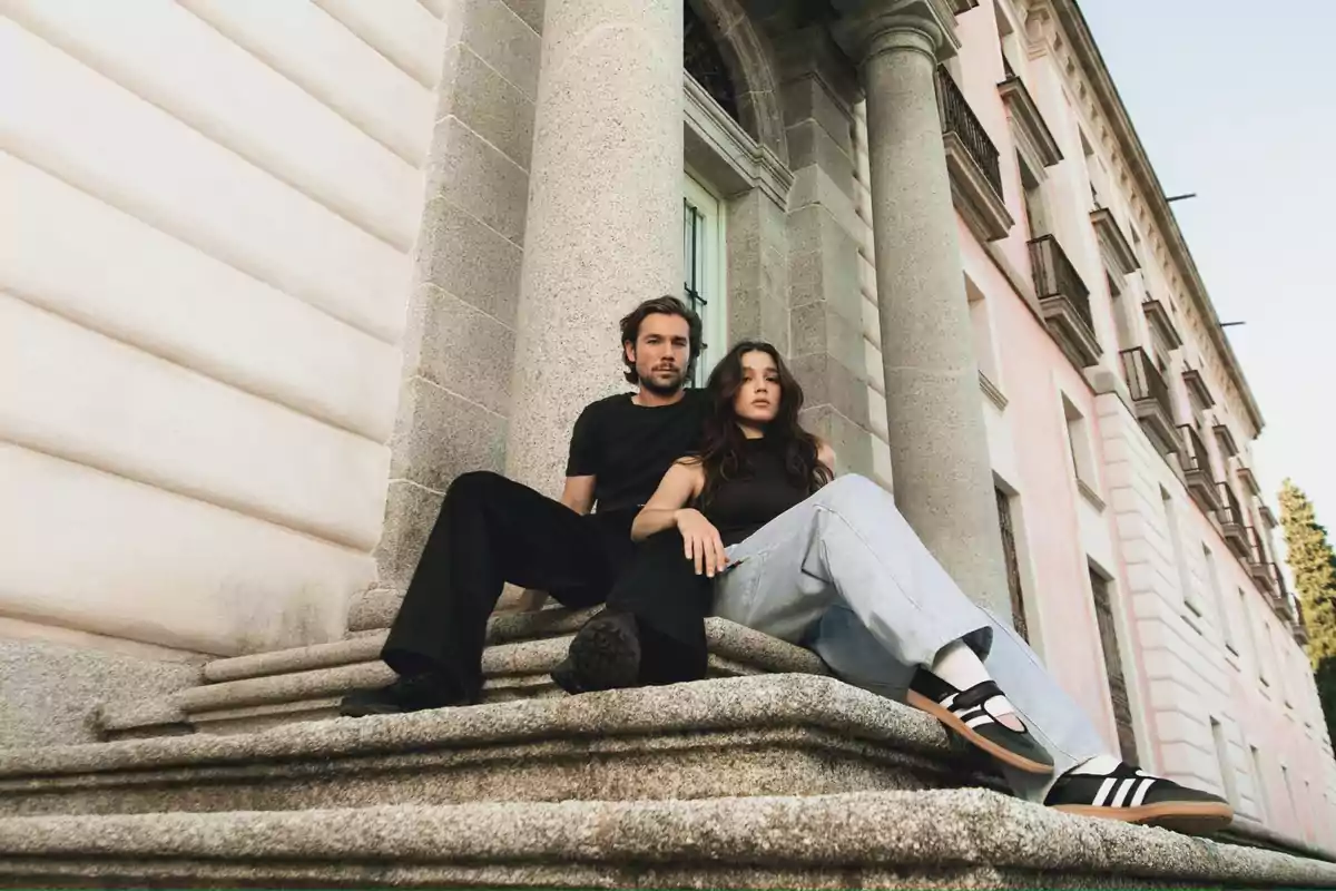 Dos personas jóvenes sentadas en las escaleras de un edificio antiguo con columnas de piedra, ambos con ropa casual y expresión seria Dos personas jóvenes sentadas en las escaleras de un edificio antiguo con columnas de piedra, ambos con ropa casual y expresión seria