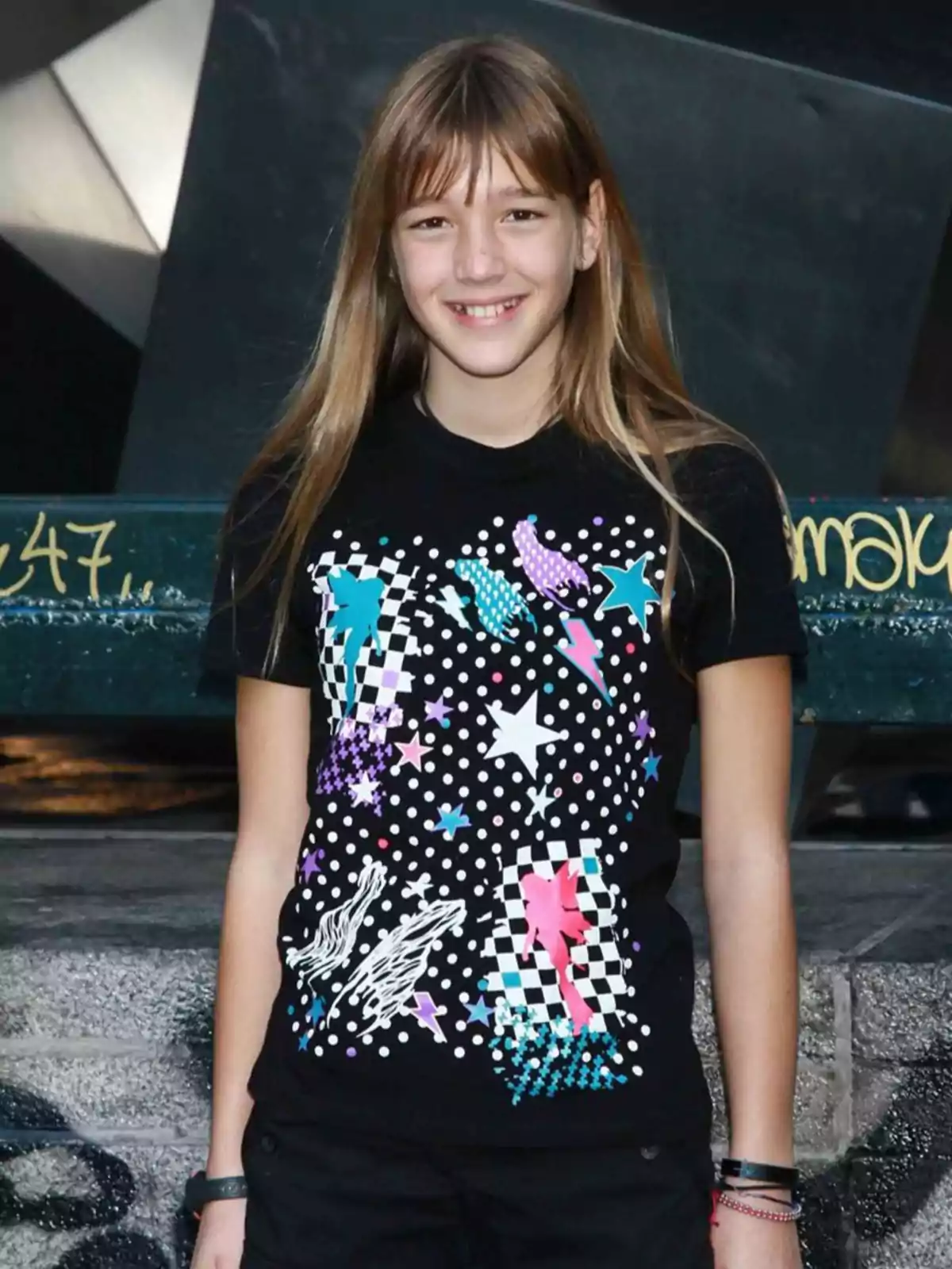 Una niña sonriente con cabello largo y lacio lleva puesta una camiseta negra con un diseño colorido de estrellas, cuadros y puntos blancos, posando al aire libre frente a una estructura urbana Una niña sonriente con cabello largo y lacio lleva puesta una camiseta negra con un diseño colorido de estrellas, cuadros y puntos blancos, posando al aire libre frente a una estructura urbana