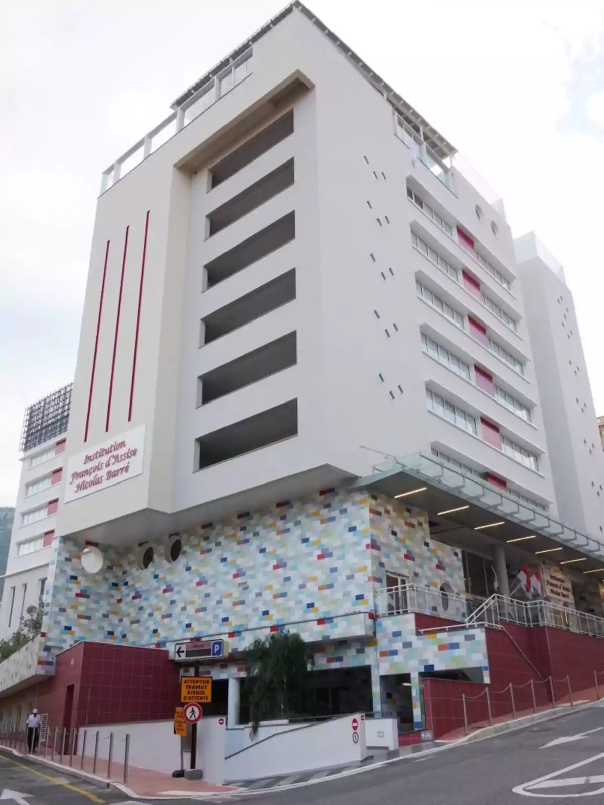 Edificio moderno de varios pisos con fachada blanca y detalles de mosaicos de colores en la planta baja, ubicado en una calle inclinada Edificio moderno de varios pisos con fachada blanca y detalles de mosaicos de colores en la planta baja, ubicado en una calle inclinada