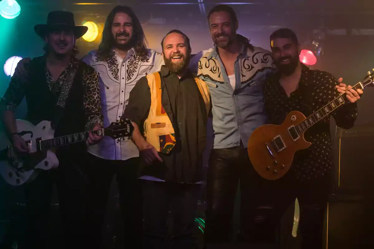 Cinco hombres sonríen en el escenario, dos de ellos sostienen guitarras eléctricas y todos visten ropa de estilo country o rock, con luces de colores al fondo. Cinco hombres sonríen en el escenario, dos de ellos sostienen guitarras eléctricas y todos visten ropa de estilo country o rock, con luces de colores al fondo.