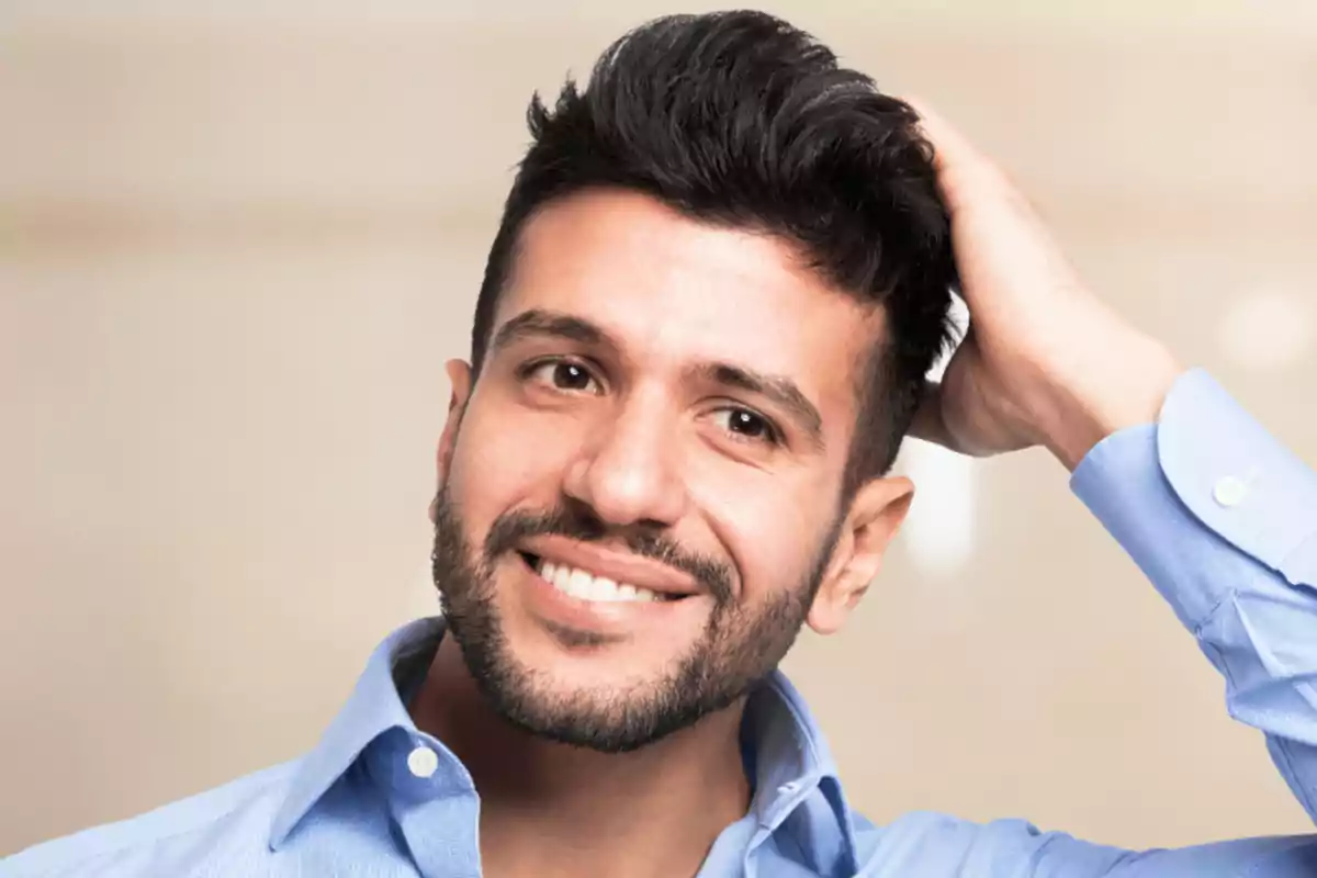 Hombre joven con barba y cabello oscuro sonriendo mientras se toca la cabeza con una mano