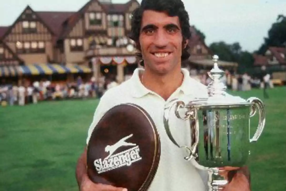 Hombre sonriente con camiseta blanca sostiene un trofeo plateado y una funda de raqueta de tenis en un campo de césped frente a un edificio antiguo Hombre sonriente con camiseta blanca sostiene un trofeo plateado y una funda de raqueta de tenis en un campo de césped frente a un edificio antiguo