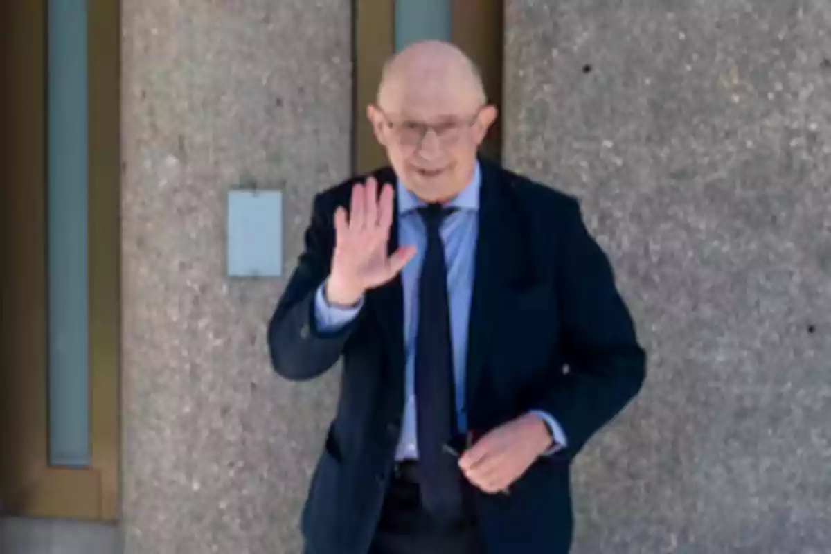 Hombre mayor con gafas y traje oscuro saludando con la mano frente a una pared de concreto Hombre mayor con gafas y traje oscuro saludando con la mano frente a una pared de concreto