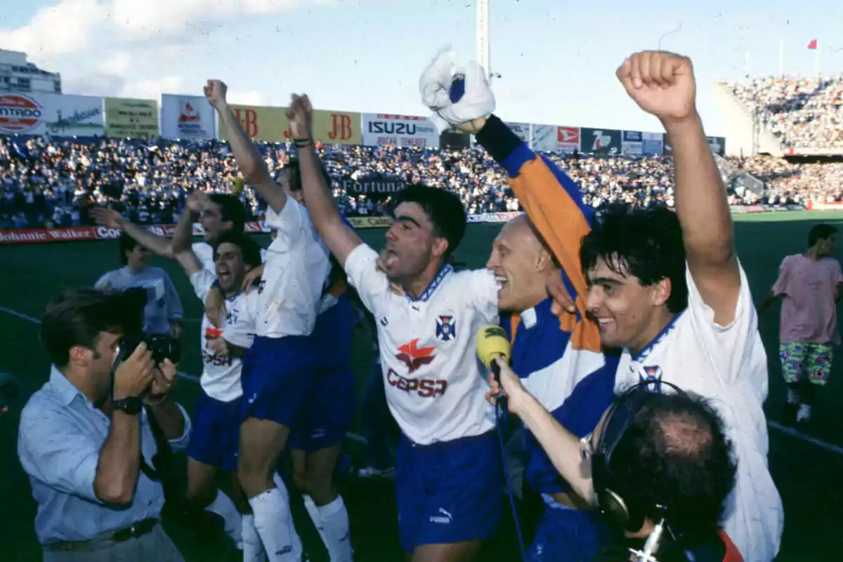 Jugadores de fútbol celebran efusivamente en el campo mientras son fotografiados y entrevistados por la prensa con el estadio lleno de aficionados al fondo Jugadores de fútbol celebran efusivamente en el campo mientras son fotografiados y entrevistados por la prensa con el estadio lleno de aficionados al fondo