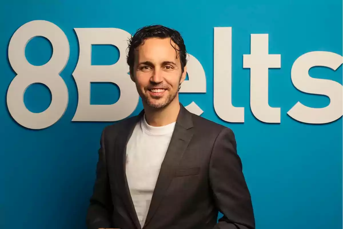 Hombre con barba corta y cabello oscuro sonríe frente a un fondo azul con el logo de 8Belts