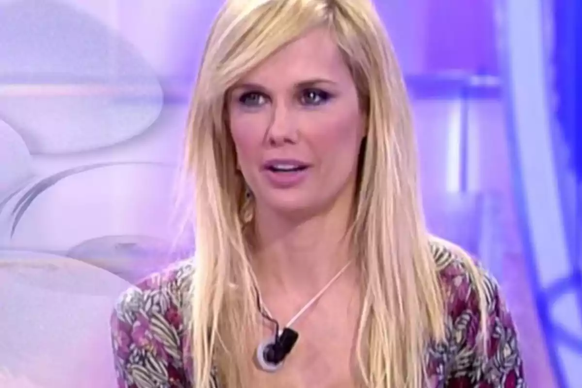 Mujer rubia con cabello largo y liso usando un micrófono de solapa en un set de televisión con fondo violeta