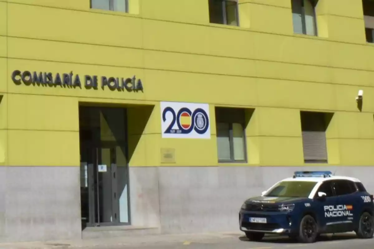 Fachada de una comisaría de policía de color amarillo con un coche de la Policía Nacional aparcado frente a la entrada y un cartel conmemorativo del bicentenario en la pared