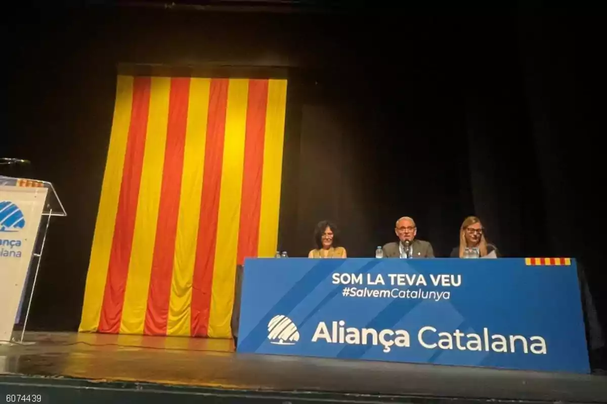 Tres personas sentadas en una mesa azul con el logo de Aliança Catalana y un gran estandarte con franjas rojas y amarillas al fondo en un escenario