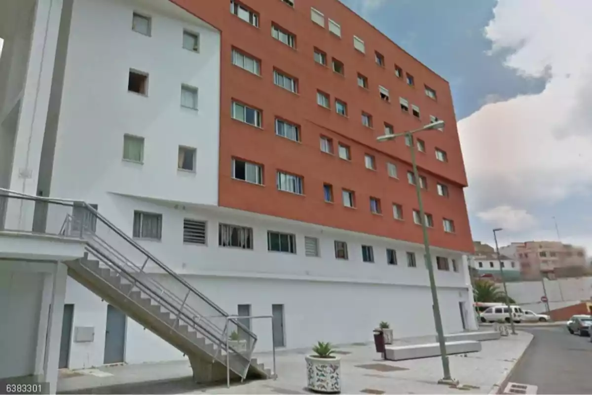 Edificio moderno de varios pisos con fachada blanca y roja, ventanas alineadas, una escalera exterior metálica y una calle con aceras y macetas en primer plano Edificio moderno de varios pisos con fachada blanca y roja, ventanas alineadas, una escalera exterior metálica y una calle con aceras y macetas en primer plano