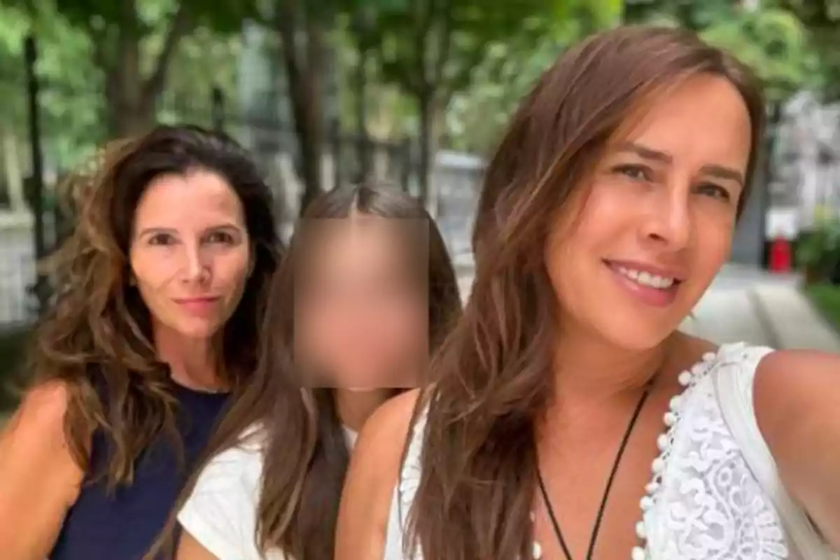Tres personas posan para una selfie al aire libre, dos mujeres adultas sonríen y una niña en el centro tiene el rostro difuminado, con árboles y un camino de fondo.